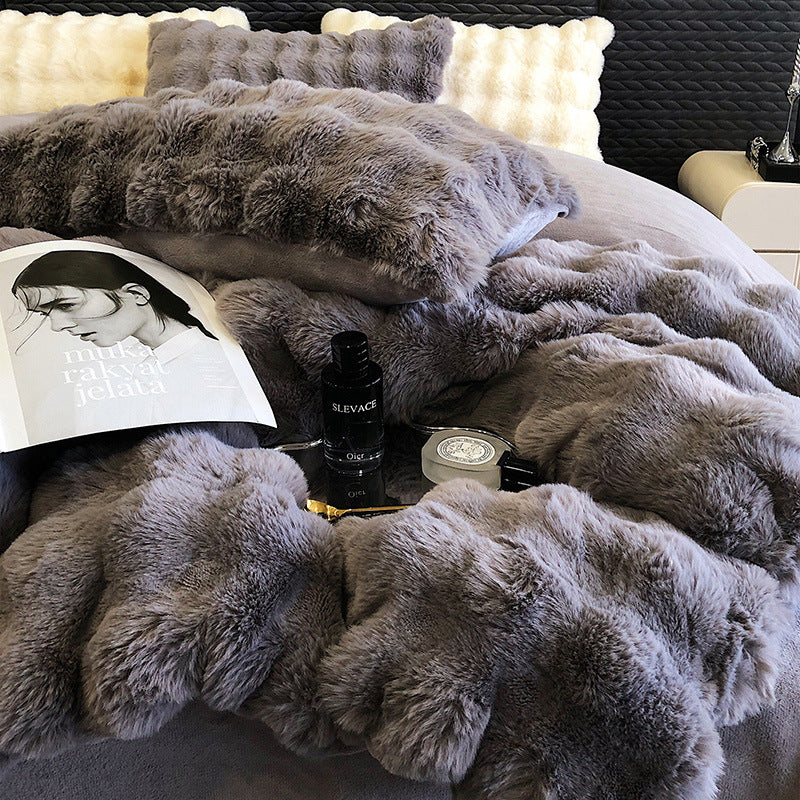 Gray faux rabbit fur beddubg draped over a bed with a book and makeup items on top.