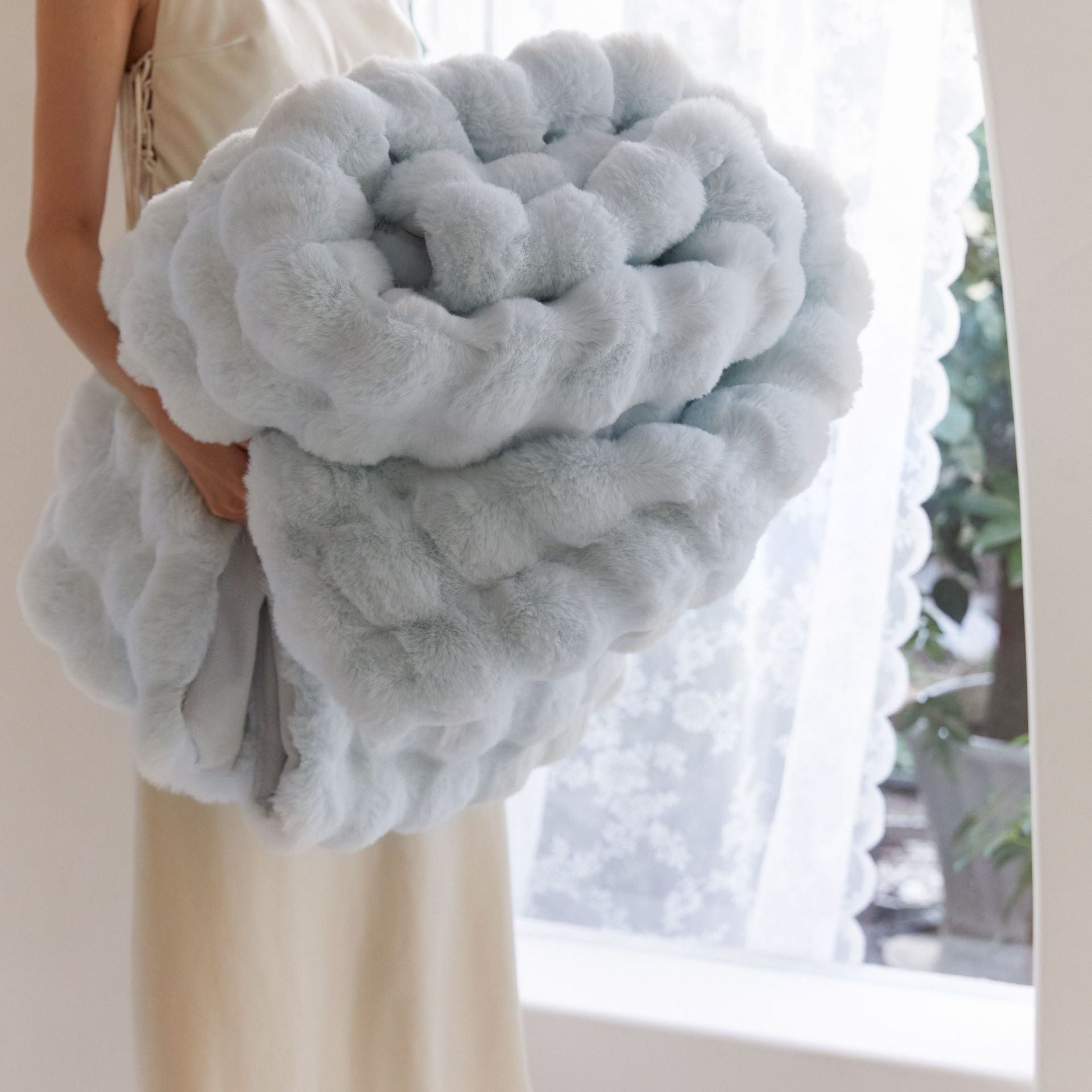 Person holding a large fluffy gray pillow in front of a window