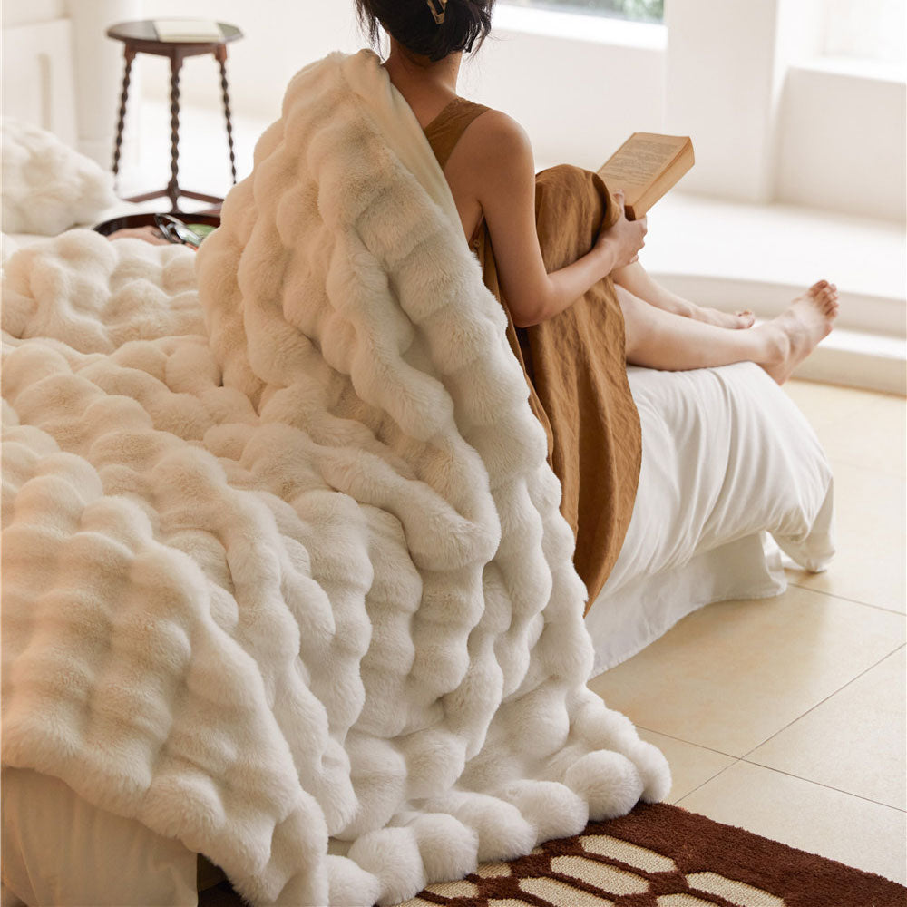 Person sitting on a bed with a fluffy white blanket and a book, in a bright room.