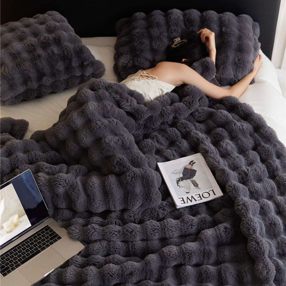 Person lying on a bed with a dark gray fluffy blanket, using a laptop.