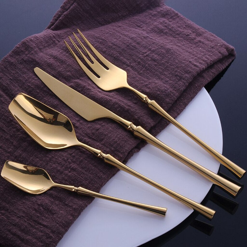Set of gold cutlery including a knife, fork, and two spoons on a white plate with a purple cloth background.