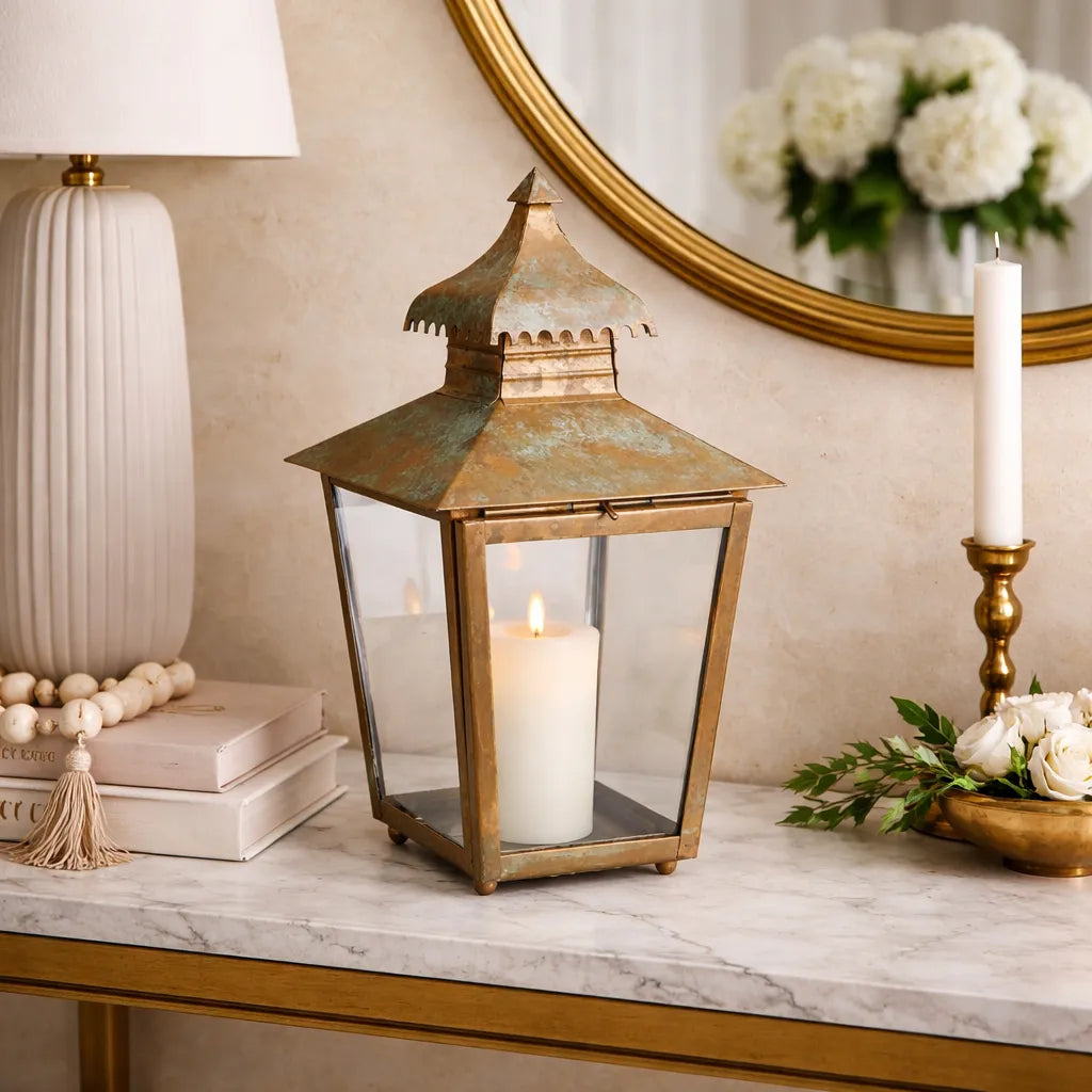 Decorative copper pagoda lantern with weathered patina and a candle on a marble surface, surrounded by books, a lamp, and flowers.