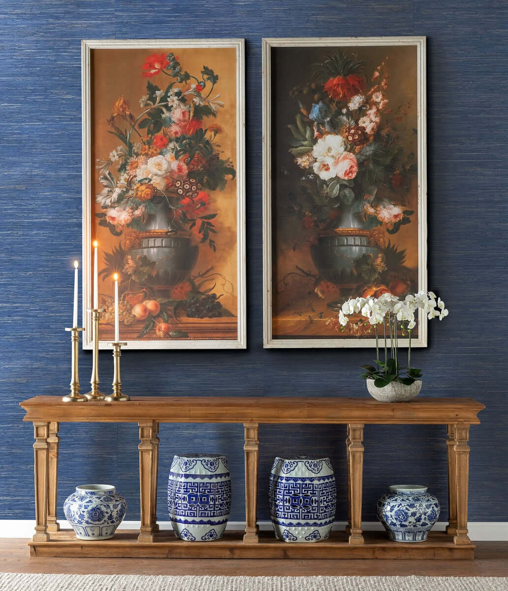 Two floral paintings above a wooden console table with decorative vases against a blue wall.