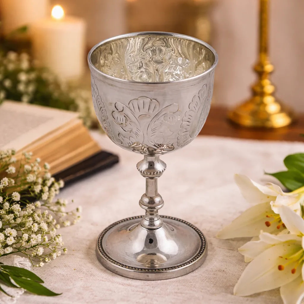 Silver plated chalice goblet with floral patterns on a table in a religious setting with flowers and a candle in the background