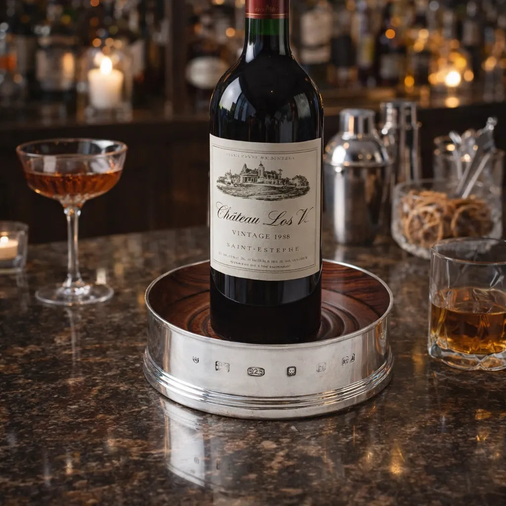 Bottle of Chateau Les V on a sterling silver coaster with mahogany wood inlay with cocktails in the background
