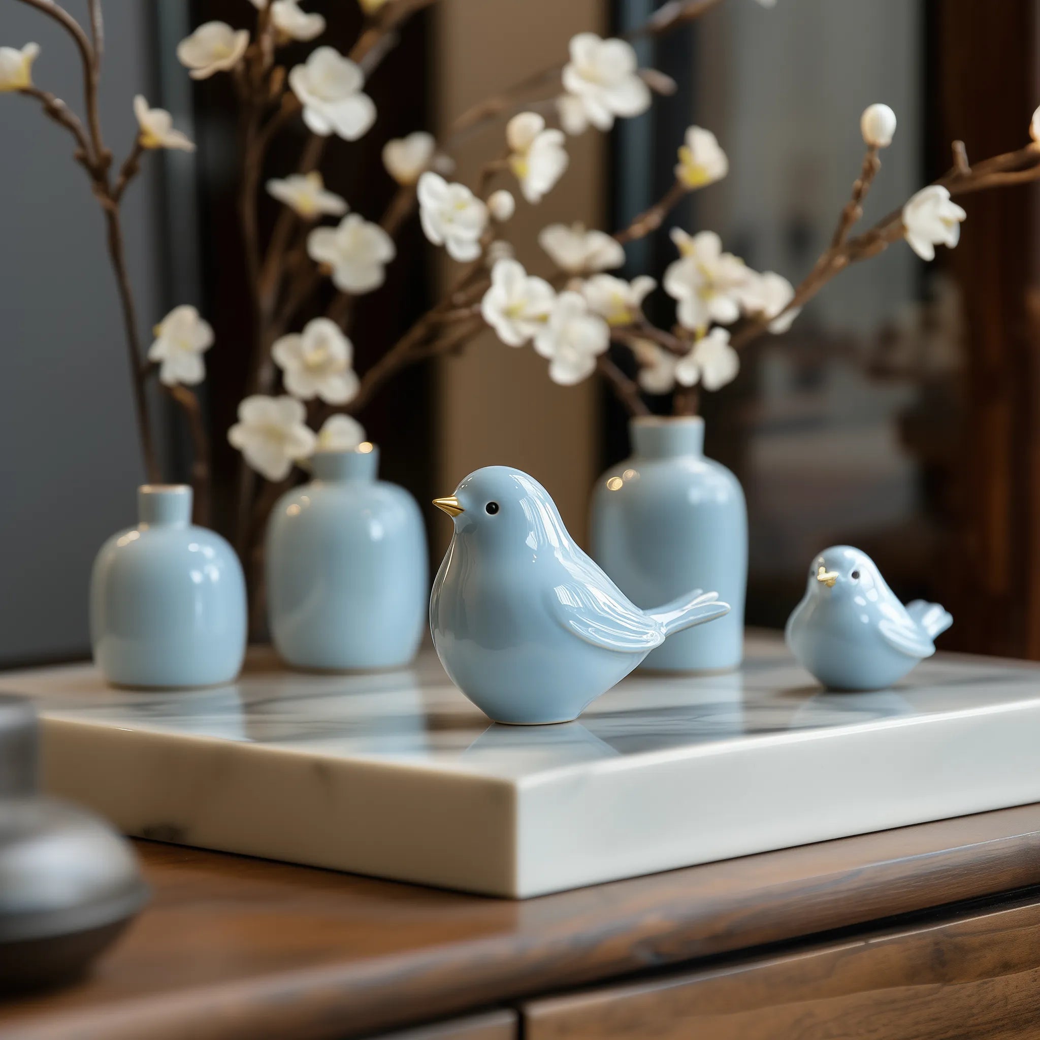 Two ceramic birds on a wooden surface with decorative vases and branches in the background.