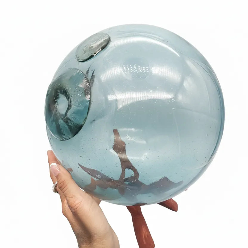 Hand holding a large rare original Japanese turquoise hand-blown glass fishing float sphere with wooden stand on a white background.