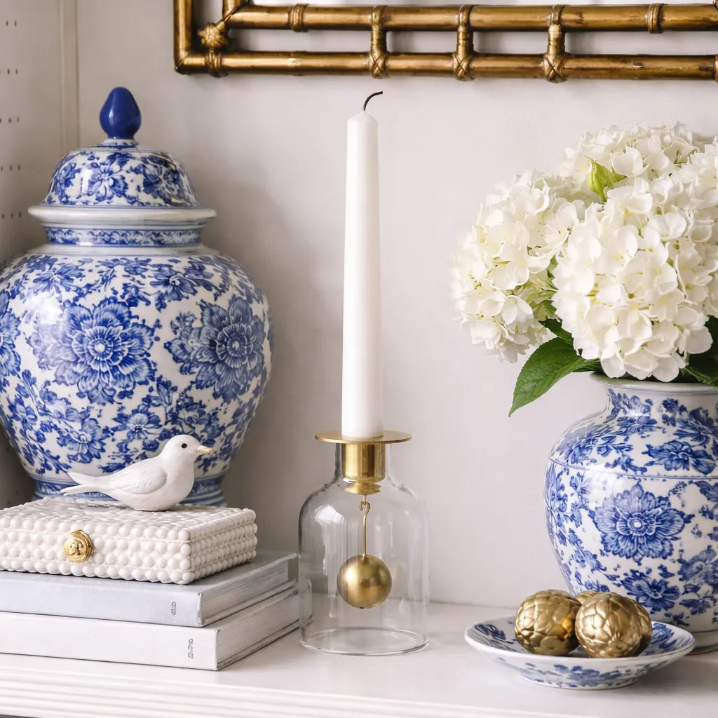 Klong Bell candlestick in glass and matte brass styled on a white shelf surrounded by blue and white chinoiserie ginger jars, hydrangeas, stacked books, and a gold bamboo mirror.