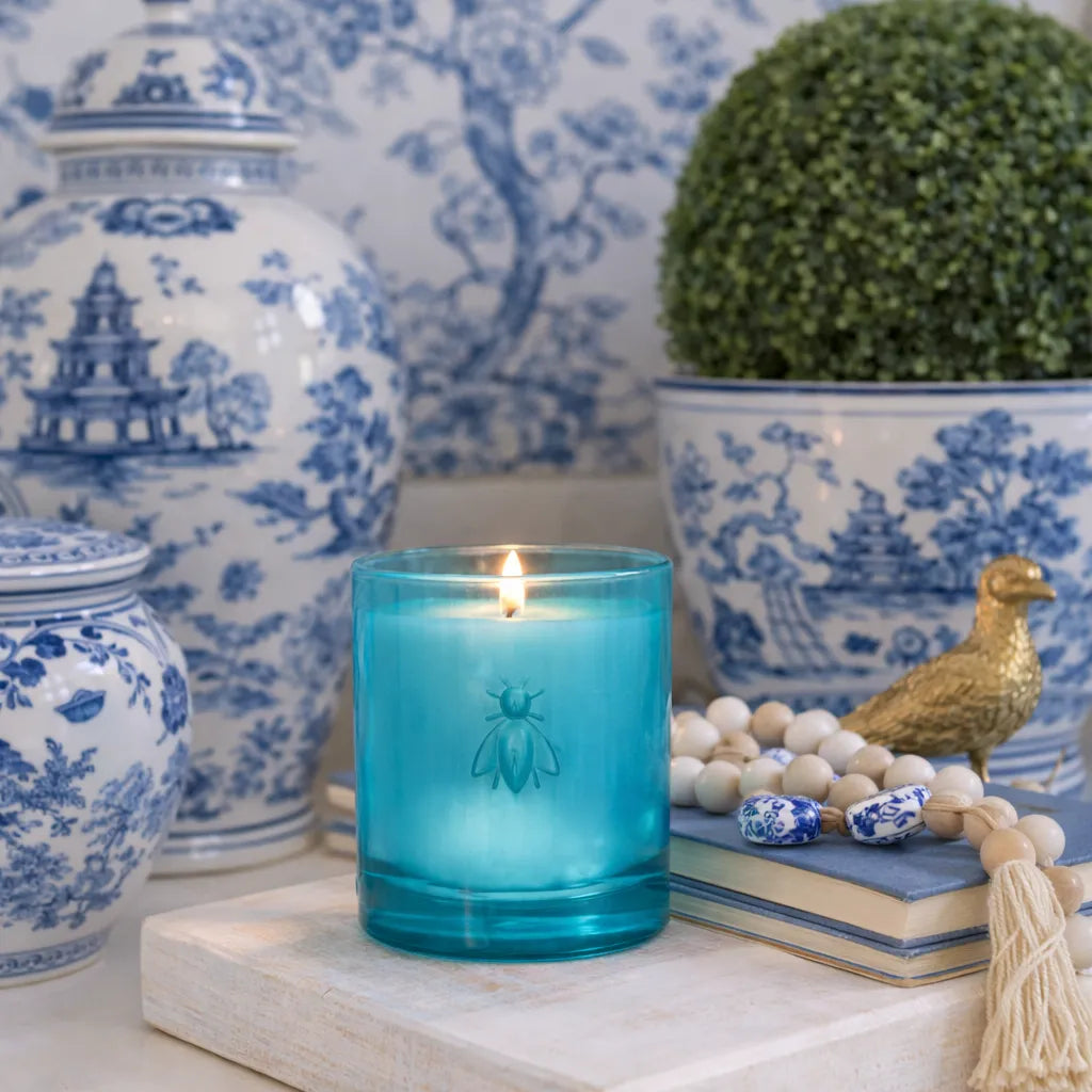 La Rochère candle in turquoise glass with a bee emblem in a decorative chinoiserie setting with blue and white jars and a potted plant.