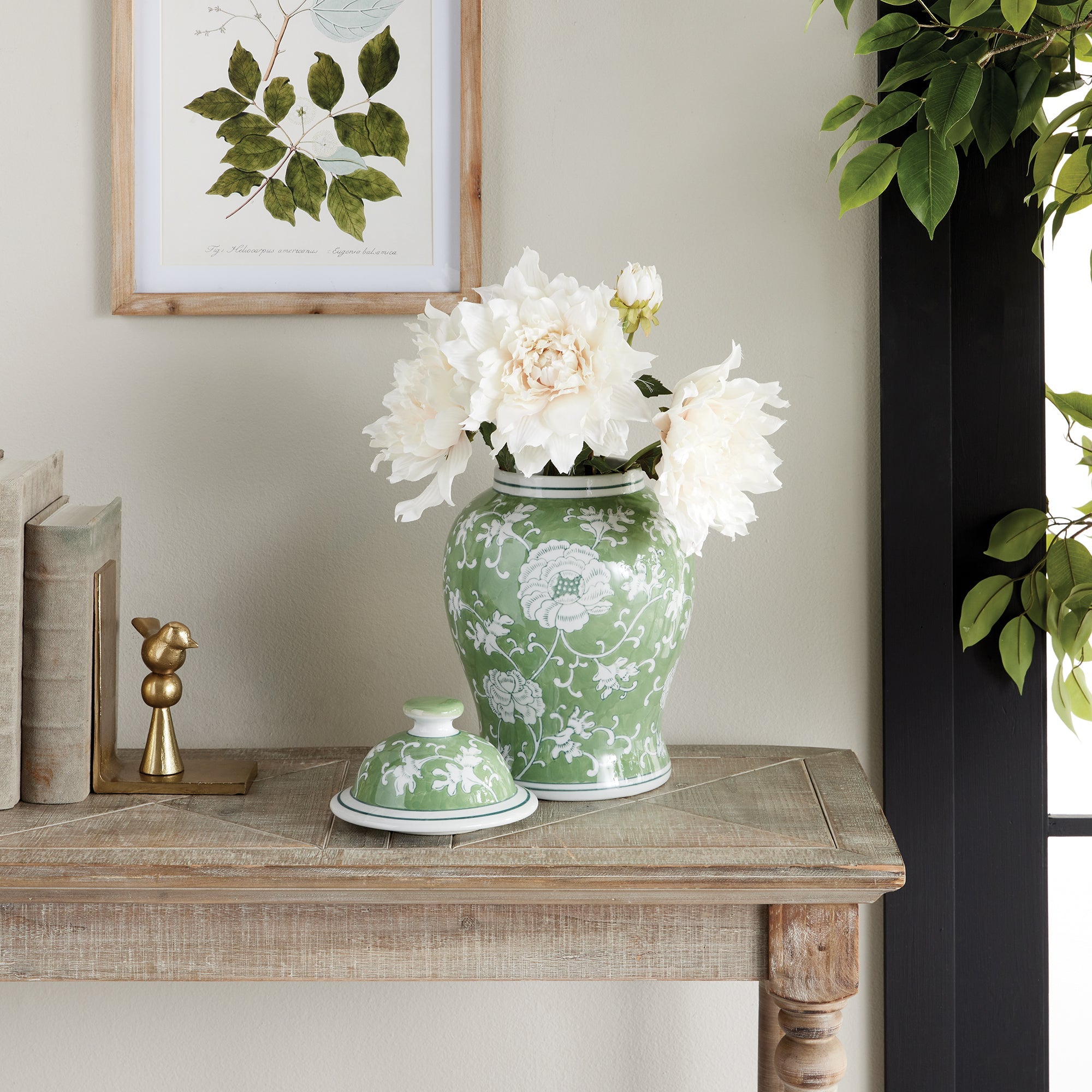 Green floral-patterned vase with white flowers on a wooden table, framed artwork in the background.