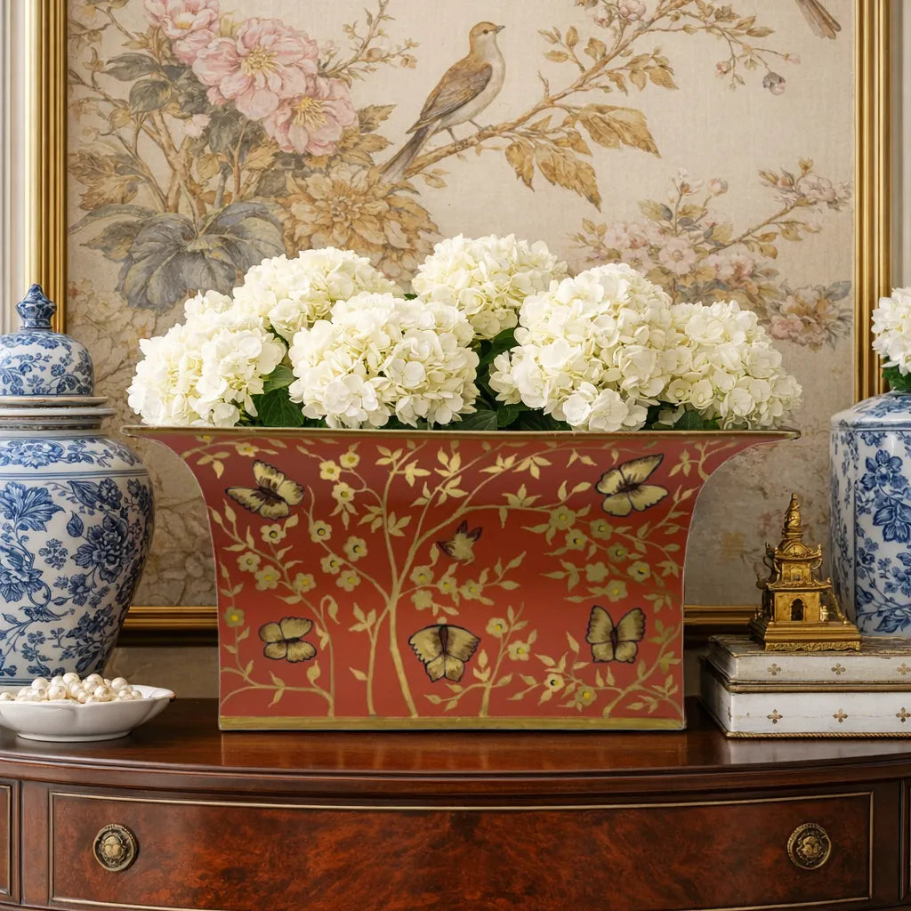 Decorative red and gold butterfly chinoiserie floral planter with white hydrangeas on a wooden table.