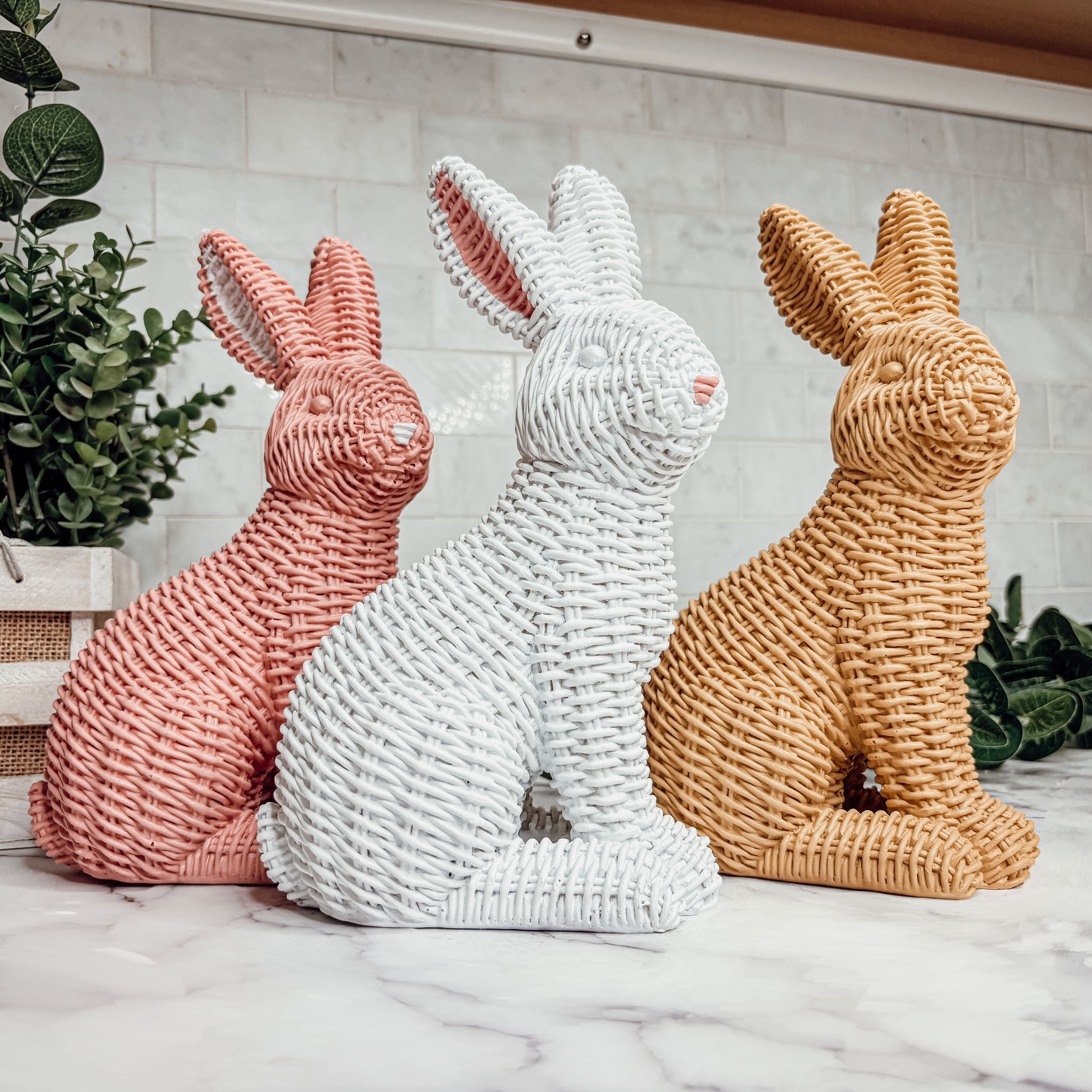 Three woven rabbit sculptures in pink, white, and brown on a marble surface with a plant in the background.