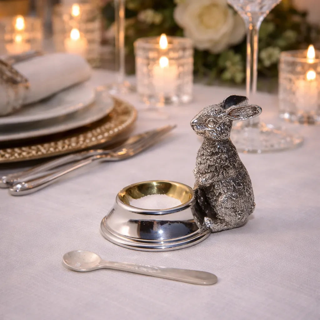 Decorative silver rabbit-shaped salt cellar and spoon on a table with candles and plates