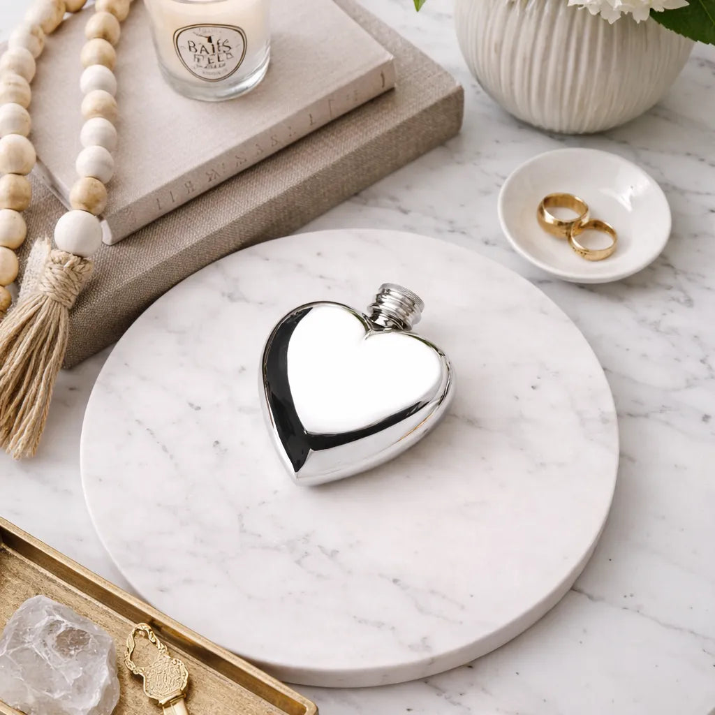 Heart-shaped pewter flask on a marble surface with jewelry and a candle in the background
