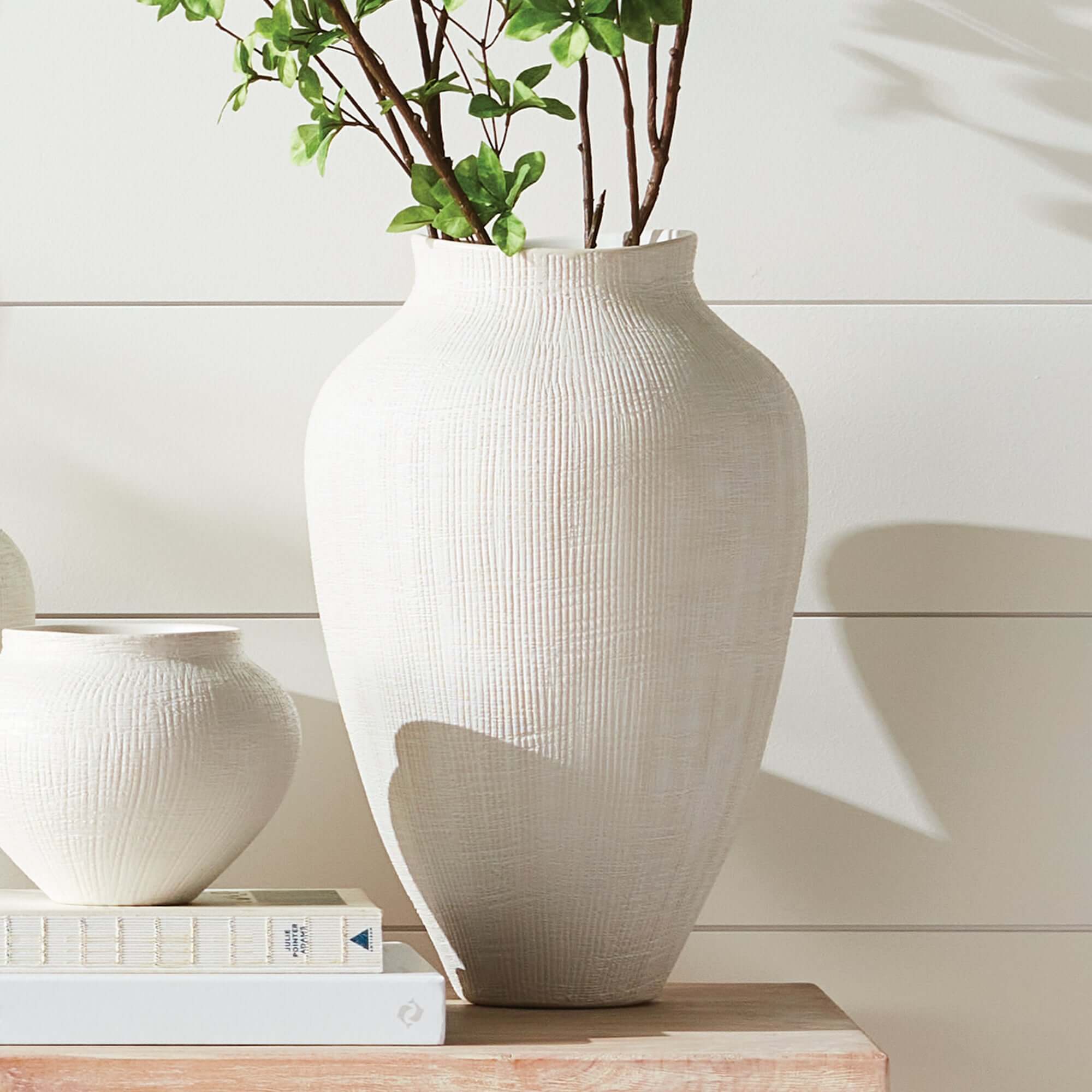 White textured vase with green plant on a wooden surface against a white wall